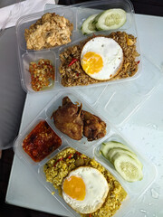Delicious Indonesian rice dishes in practical take-out containers, complete with fried eggs, chicken, and fresh cucumber slices.