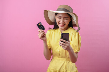 Asian woman wearing a summer hat and yellow dress smiling while holding a credit card and...