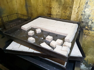 Freshly made tofu, some cut into cubes, rests on large trays in a production setting with a rustic,...