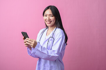 Asian female doctor using a smartphone with both hands while smiling, dressed in a lab coat and...