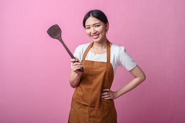 Asian woman wearing a brown apron smiling confidently while holding a long wooden spatula, standing...