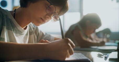 Elementary School Boy Writing Exam or Doing Task in Notebook. Female Teacher Walking Between Desks, Controlling Young Boys and Girls During School Test Process in Modern Classroom. Close Up Shot.