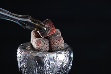 Putting smoldering charcoal cube onto hookah holder covered with foil on black background, closeup
