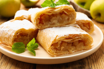 Pieces of tasty apple strudel with powdered sugar, mint and fruits on wooden table, closeup