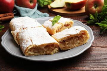 Pieces of tasty apple strudel with powdered sugar, mint and fruits on wooden table, closeup