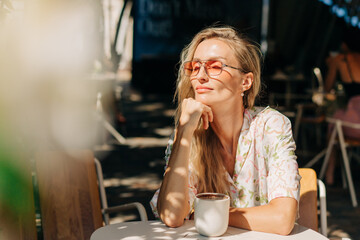 A beautiful blonde woman with long wavy hair sits on the terrace of a cafe on a summer day.