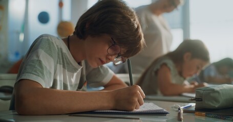 Primary School Boy Writing School Test, Doing Task in Notebook. Female Teacher Walking Between Desks, Controlling Group of Diverse Kids During the Exam Process in Modern Classroom. Close Up Shot.