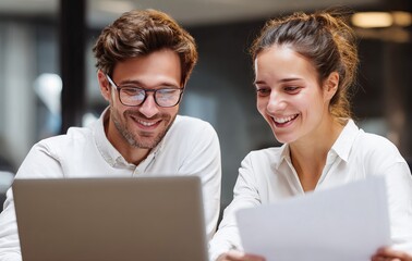  young man and woman are sitting at a desk, using a laptop computer while looking happy in an office. The man, wearing glasses, is holding a pen and pointing at the screen of a MacBook Pro