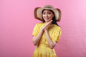 Asian woman in a yellow dress and straw hat smiles sweetly with hands resting under chin, creating a cute and peaceful expression. Shot against a vibrant pink background in studio lighting
