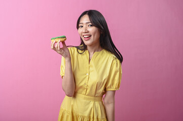 Asian woman in a yellow dress about to bite a green-sprinkled donut while looking at the camera,...
