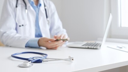 Closeup of blue stethoscope is lying on the table near female doctor checking smartphone...
