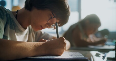 Elementary School Boy Writing Exam or Doing Task in Notebook. Female Teacher Walking Between Desks,...