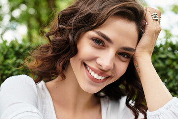 Young woman with wavy brown hair smiles in a sunny park filled with greenery