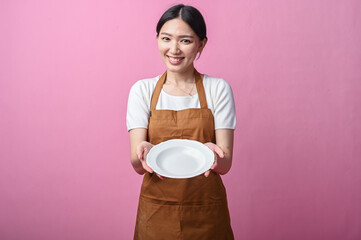 Asian woman in a brown apron holding a clean white ceramic plate with both hands, smiling warmly...