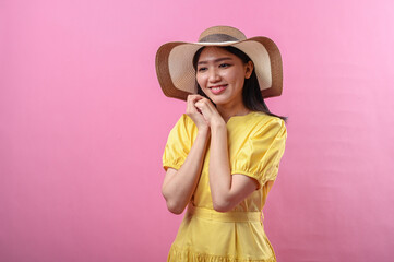 Asian woman in a yellow dress and straw hat smiles sweetly with hands resting under chin, creating...