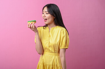 Asian woman in a yellow dress about to bite a green-sprinkled donut while looking at the camera,...