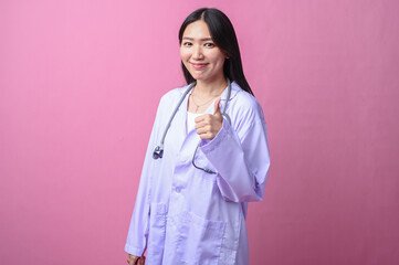 Asian female doctor smiling while pointing sideways with her thumb, wearing a stethoscope and white...