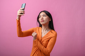 Asian woman with long black hair wearing orange dress pointing upward while holding smartphone in...