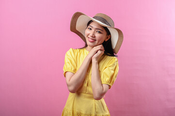 Naklejka premium Asian woman in a yellow dress and straw hat smiles sweetly with hands resting under chin, creating a cute and peaceful expression. Shot against a vibrant pink background in studio lighting