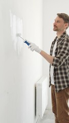 Middle aged man wearing gloves is painting a white wall with a paint roller, working on a home...
