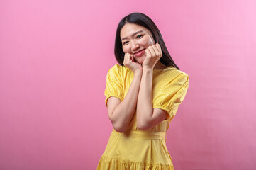 Asian woman wearing a bright yellow dress makes a cute pose with both fists gently resting on her cheeks, smiling sweetly against a soft pink background in a playful and joyful expression