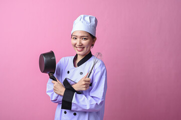 Asian female chef smiling confidently while crossing arms, holding a whisk and a saucepan, wearing...