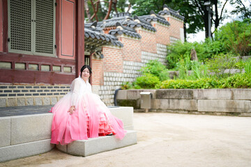 A Korean woman in her twenties wearing a charming pink hanbok sits in front of the traditional...