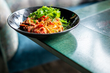 Asian noodle soup with sliced meat, herbs, and vegetables in a black bowl on green wooden table, top view. Vibrant colors and appetizing presentation.