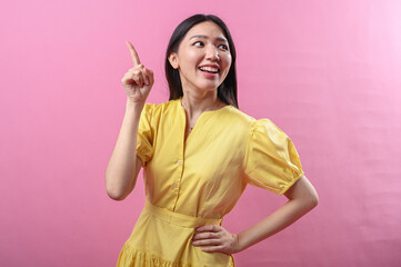 Asian woman in a yellow dress excitedly points upward with her index finger while smiling brightly....