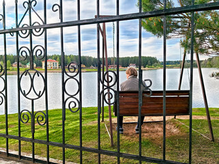 Person sitting on a swing by a calm lake surrounded by greenery and houses on a cloudy day