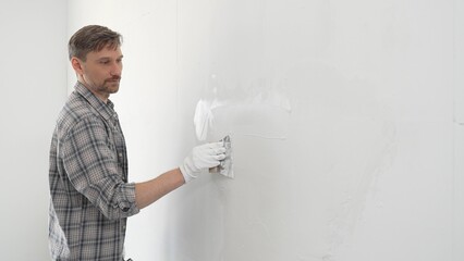 Construction worker spreading plaster on white wall, wearing protective gloves and t-shirt during...