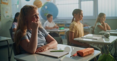 Focus on Elementary School Girl Writing in School Notebook During Geography Lesson in Modern Classroom. Team of Smart Diverse Kids Sitting at Desks, Studying Ecology, Listening to Lecture from Teacher
