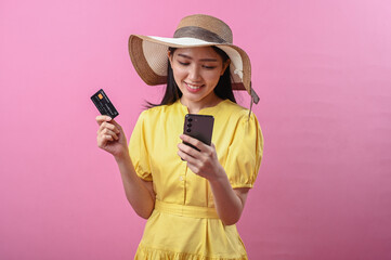 Asian woman wearing a summer hat and yellow dress smiling while holding a credit card and...