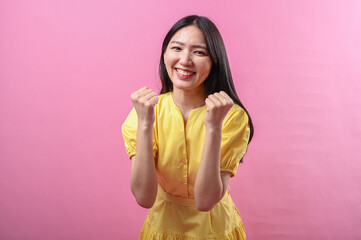Asian woman in a yellow dress smiling confidently while raising both fists in a celebratory pose....