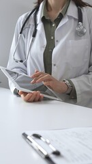 Female doctor in lab coat and stethoscope using digital tablet at desk in medical office, accessing...
