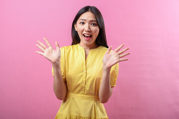 Asian woman in a yellow dress smiling brightly with both hands raised in an open wave gesture,...