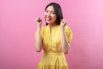 Asian woman in a yellow dress smiling confidently while raising both fists in a celebratory pose....