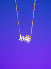 Close-up of a metallic gold pendant in the shape of "1998" on a chain, on a  purple background