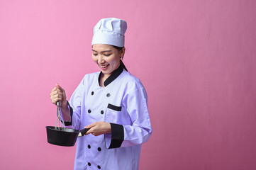 Asian female chef smiling while stirring a whisk in a small black pan, wearing a white chef uniform...