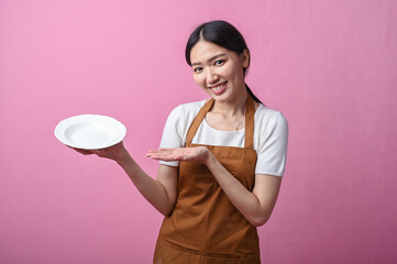 Asian woman in a brown apron smiling and pointing at an empty white plate in her hand with a pink...