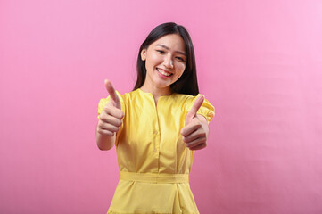 Asian woman in a yellow dress smiling with both thumbs up against a bright pink background....