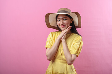 Asian woman in a yellow dress and straw hat smiles sweetly with hands resting under chin, creating...