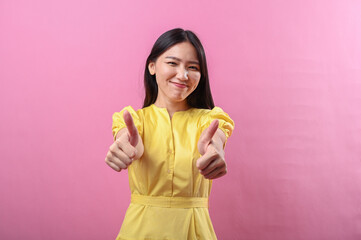 Asian woman in a yellow dress smiling with both thumbs up against a bright pink background....
