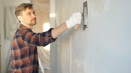 Male construction worker wearing protective gloves smoothing plaster onto wall surface during home...