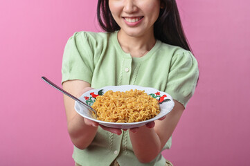 Asian woman smiling while holding a plate of cooked instant noodles with a spoon, wearing a green...