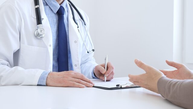 Doctor man taking notes while listening to a female patient explaining her symptoms during a medical consultation in the clinic office. Medicine and health care concept