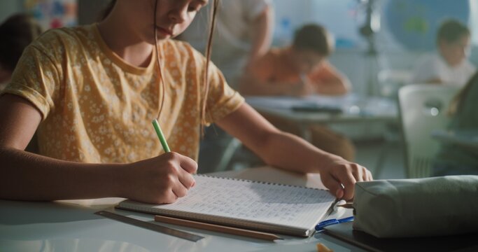 Primary School Girl Writing School Test, Doing Task in Notebook. Female Teacher Walking Between Desks, Controlling Group of Diverse Kids During the Exam Process in Modern Classroom. Close Up Shot.