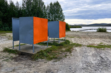 Two open air cabana in vibrant orange and grey tones, located on a sandy beach area next to a quiet...