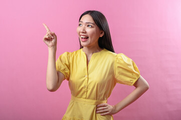 Asian woman in a yellow dress excitedly points upward with her index finger while smiling brightly....