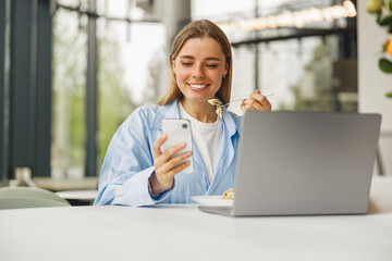 A cheerful young woman enjoys her meal while multitasking on her smartphone and laptop in a modern...